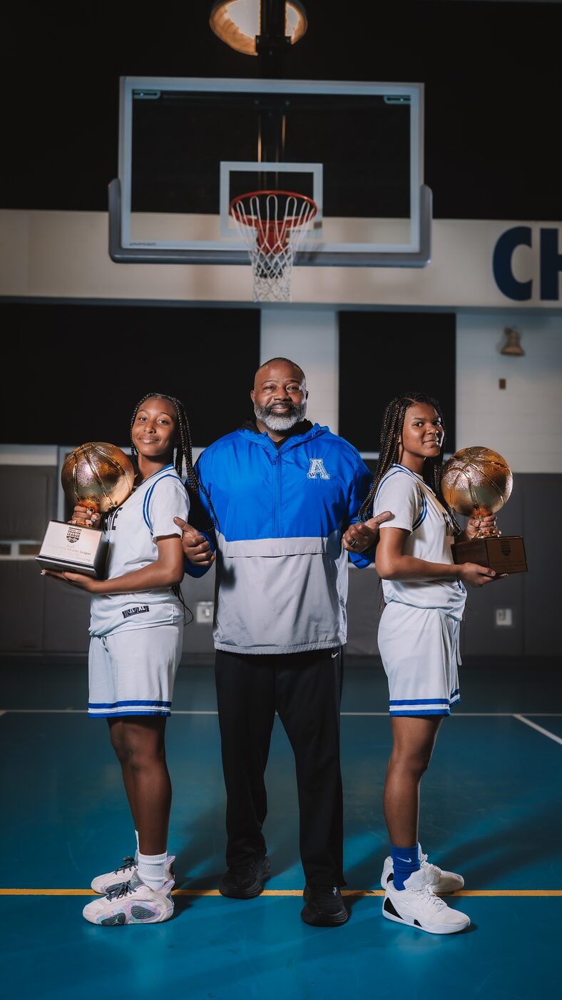 A coach stands with two players holding championship trophies under a basketball hoop.