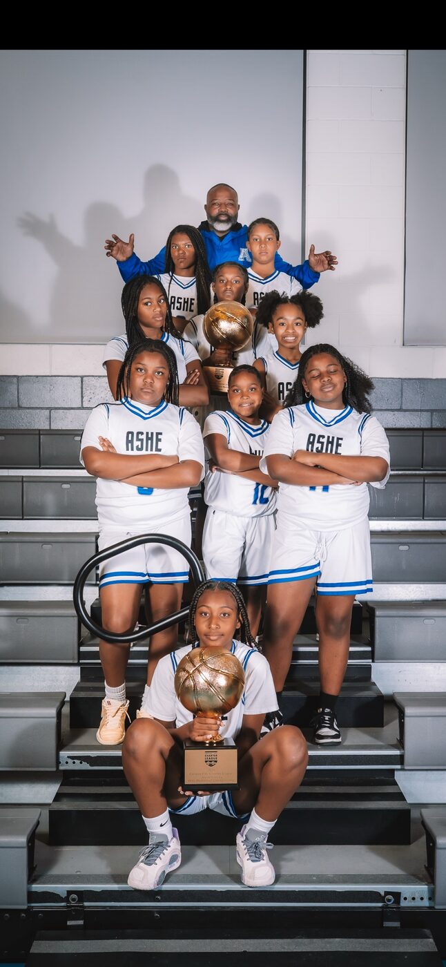 A girls basketball team poses on gym bleachers with championship trophies.