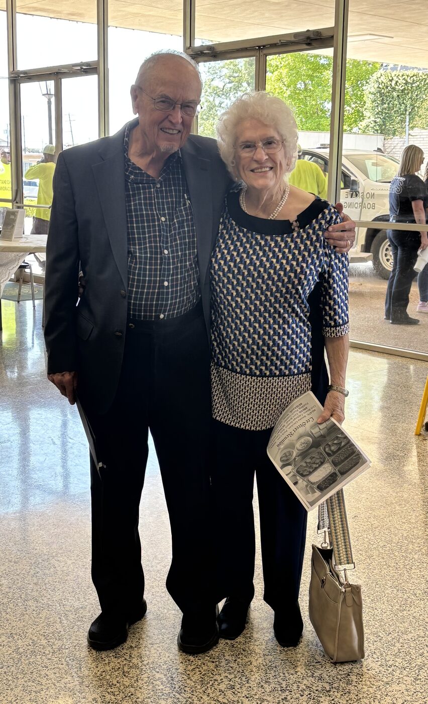 An older couple smiles together in a sunlit lobby holding an event program.