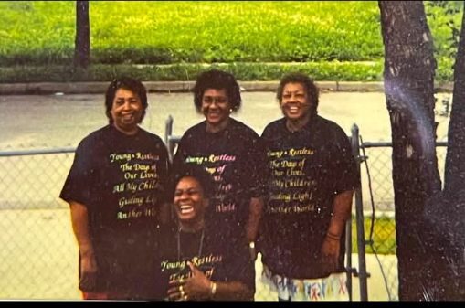 A vintage photo of four women in matching shirts posing by a chain-link fence.