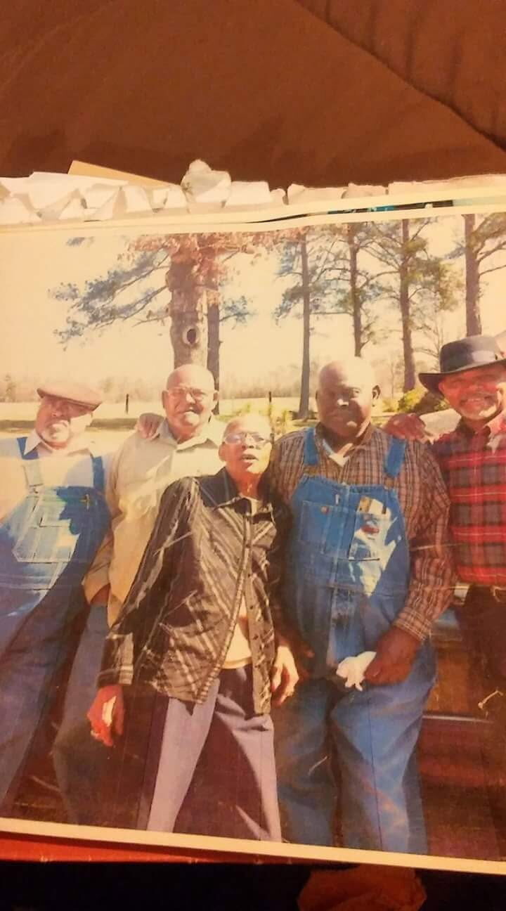 Vintage printed photo of five older men standing together outdoors under tall pine trees.
