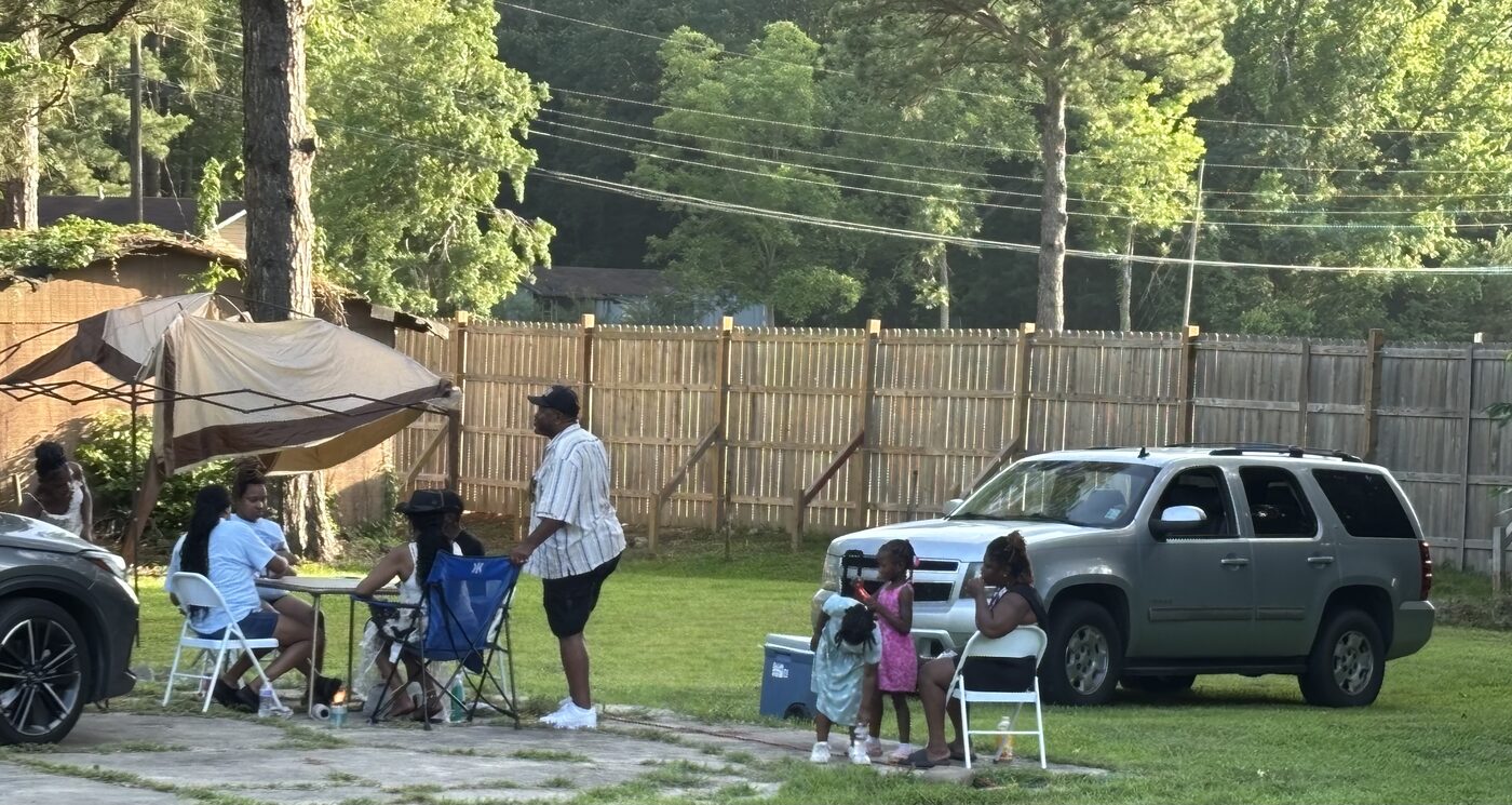 Families relax under a canopy and around folding chairs in a backyard on a sunny evening.