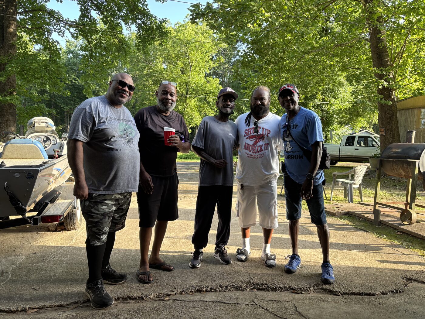 Five friends stand together on a shaded driveway with a boat and barbecue pit nearby.