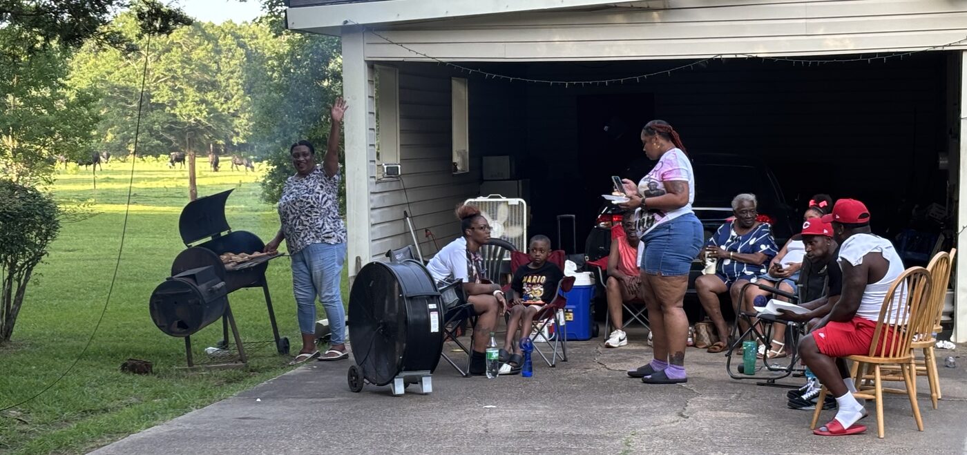 A woman waves from the barbecue pit while family members gather in the shade of a carport.