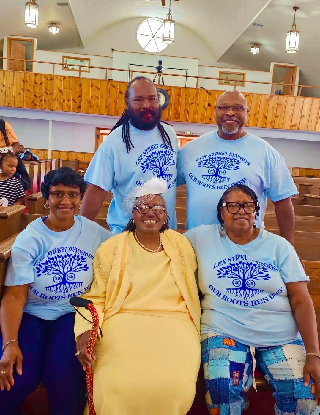 Family members in matching reunion shirts gather around a seated elder in a church sanctuary.