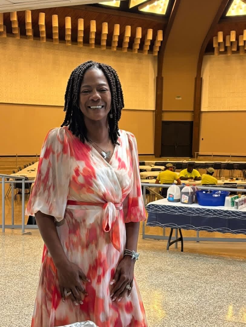 A smiling woman in a floral dress stands in a large event hall as tables are set up.