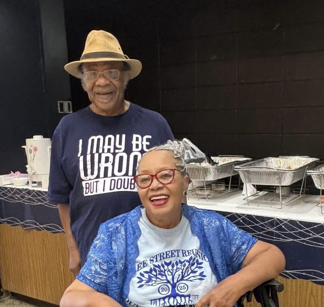 Two smiling attendees in Lee Street Reunion shirts pose near the buffet line at an indoor gathering.