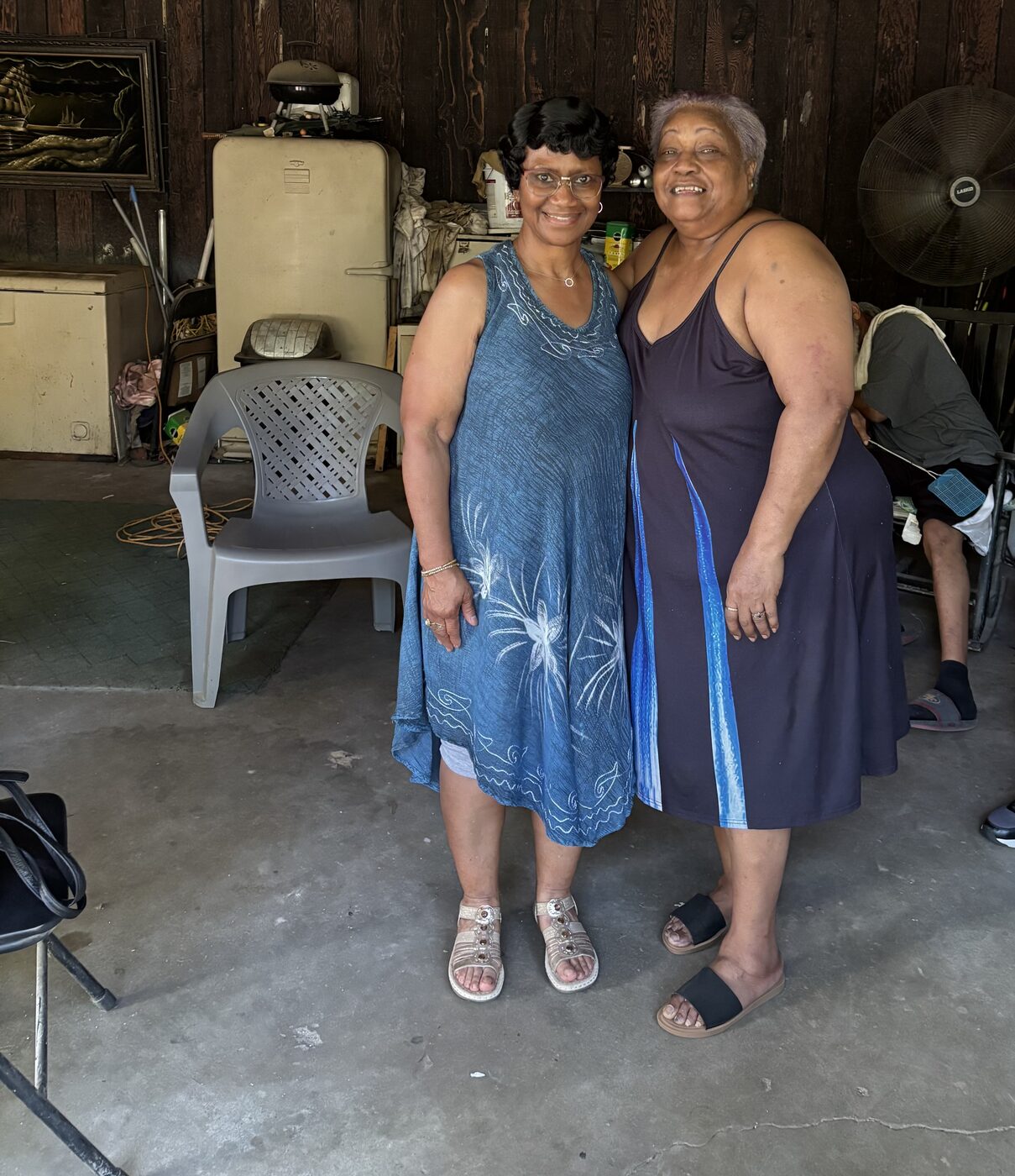 Two women in blue dresses smile side by side inside a garage during a family gathering.