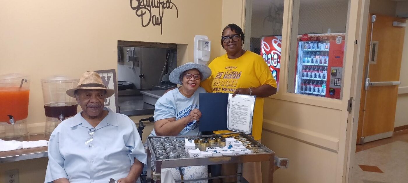 Three people in reunion shirts pose with a certificate beside a cart of party favors in a community hall.