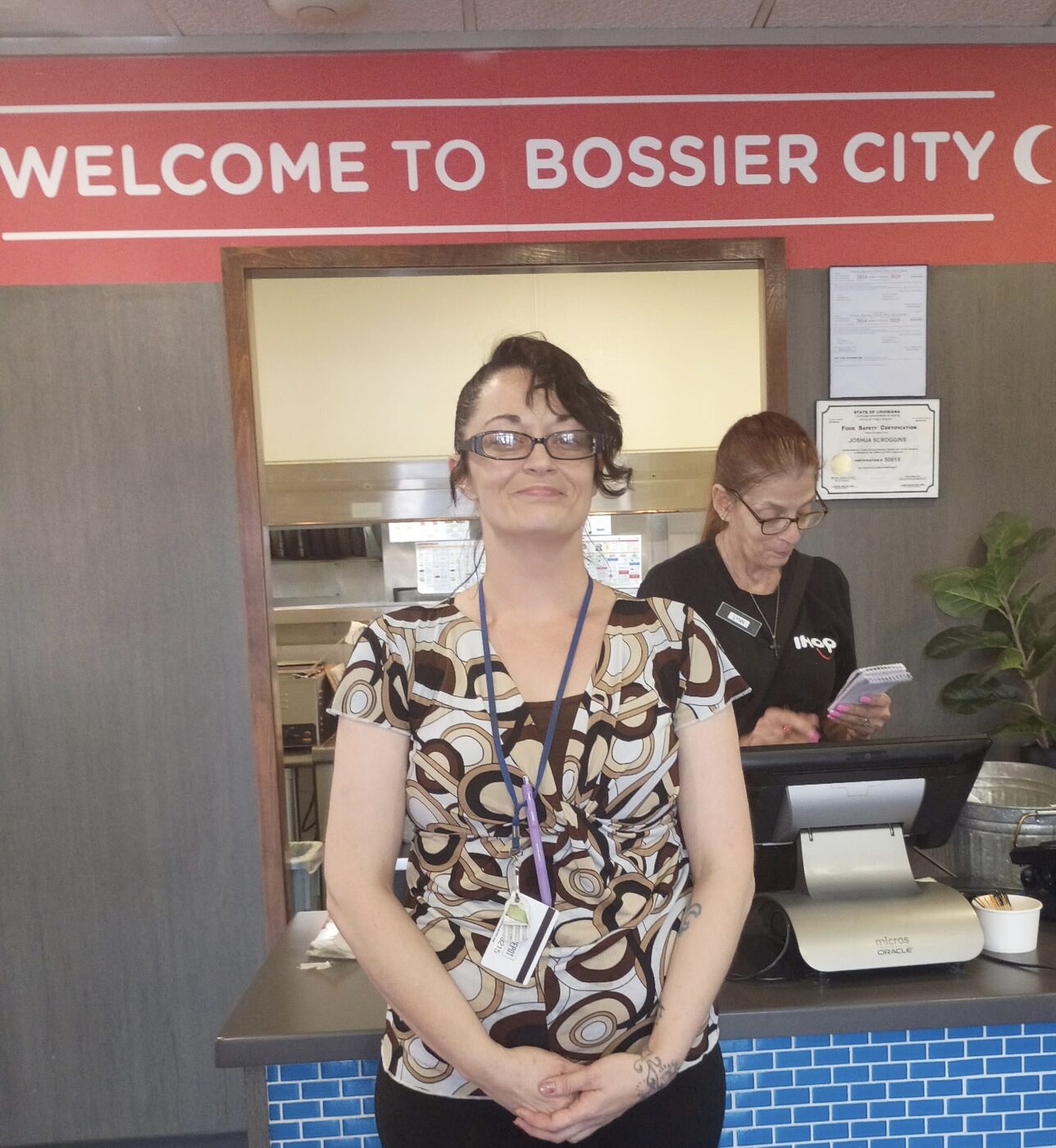 A smiling restaurant worker stands at the front counter under a welcome sign.
